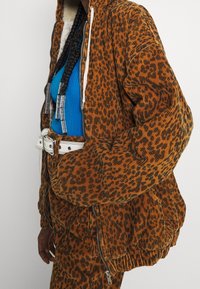 Huppuinen leopardi-nahkakuosiset corduroy-takki, jossa on etuvetoketju ja rento istuvuus. Yhdistetty siniseen ribattuun paitaan ja valkoisiin vyökorostuksiin.