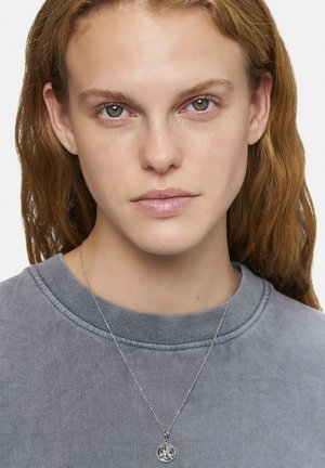 Silberne Anhängerkette mit einem runden Charm, der dekorative Details aufweist, getragen über einem grauen Sweatshirt mit strukturiertem Rundhalsausschnitt.