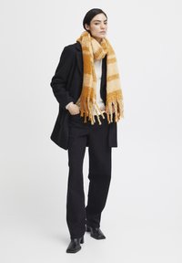 Manteau en laine noir, pull beige, pantalon noir, et écharpe rayée jaune et crème avec des détails à franges. Bottes noires à talons carrés.