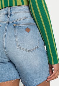 Vaaleansiniset korkeavyötäröiset farkkushortsit, joissa on repsottavat reunat ja pieni ruskea nahkalaatta takataskussa, yhdistettynä teksturoituun vihreään raitapaitaan.