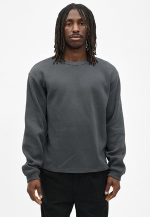Man with shoulder-length dreadlocks wearing a long-sleeve dark gray textured shirt and black pants, standing against a plain white background.