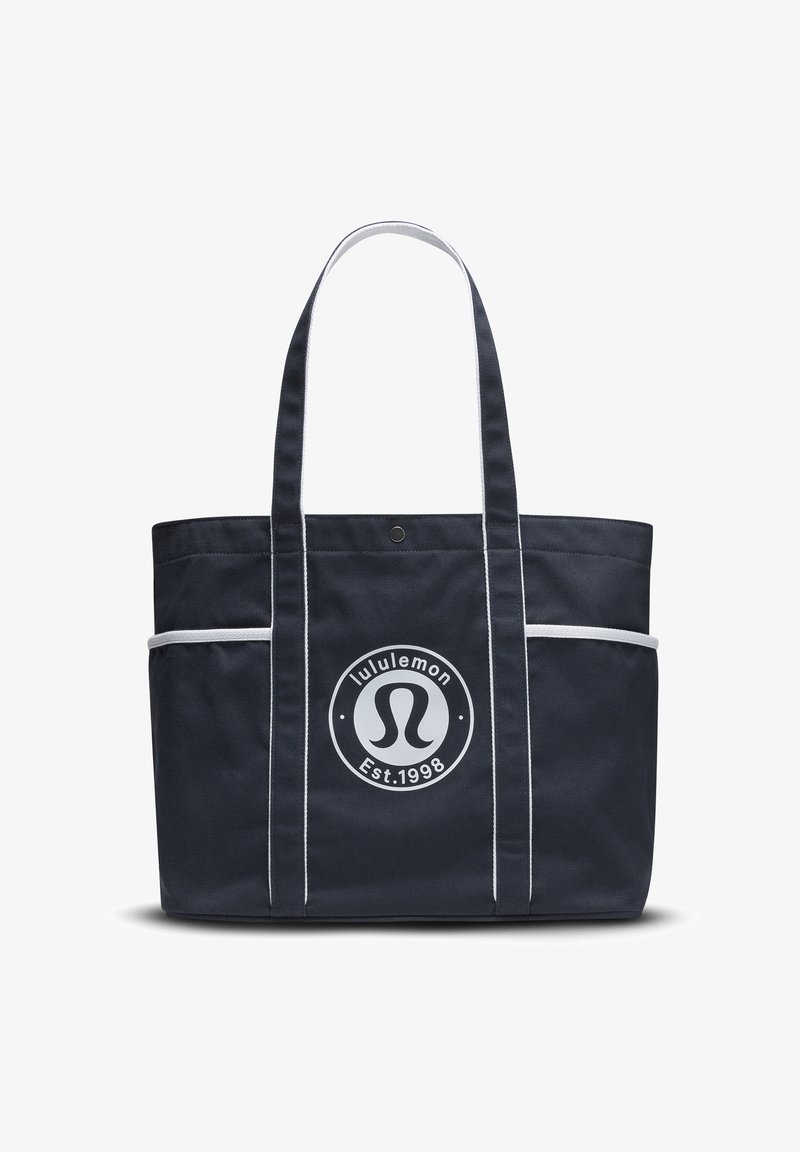 Bolsa de tote azul marino con ribetes blancos, que presenta de manera destacada un logo circular. Confeccionada con un tejido duradero, cuenta con dos asas resistentes y bolsillos laterales.