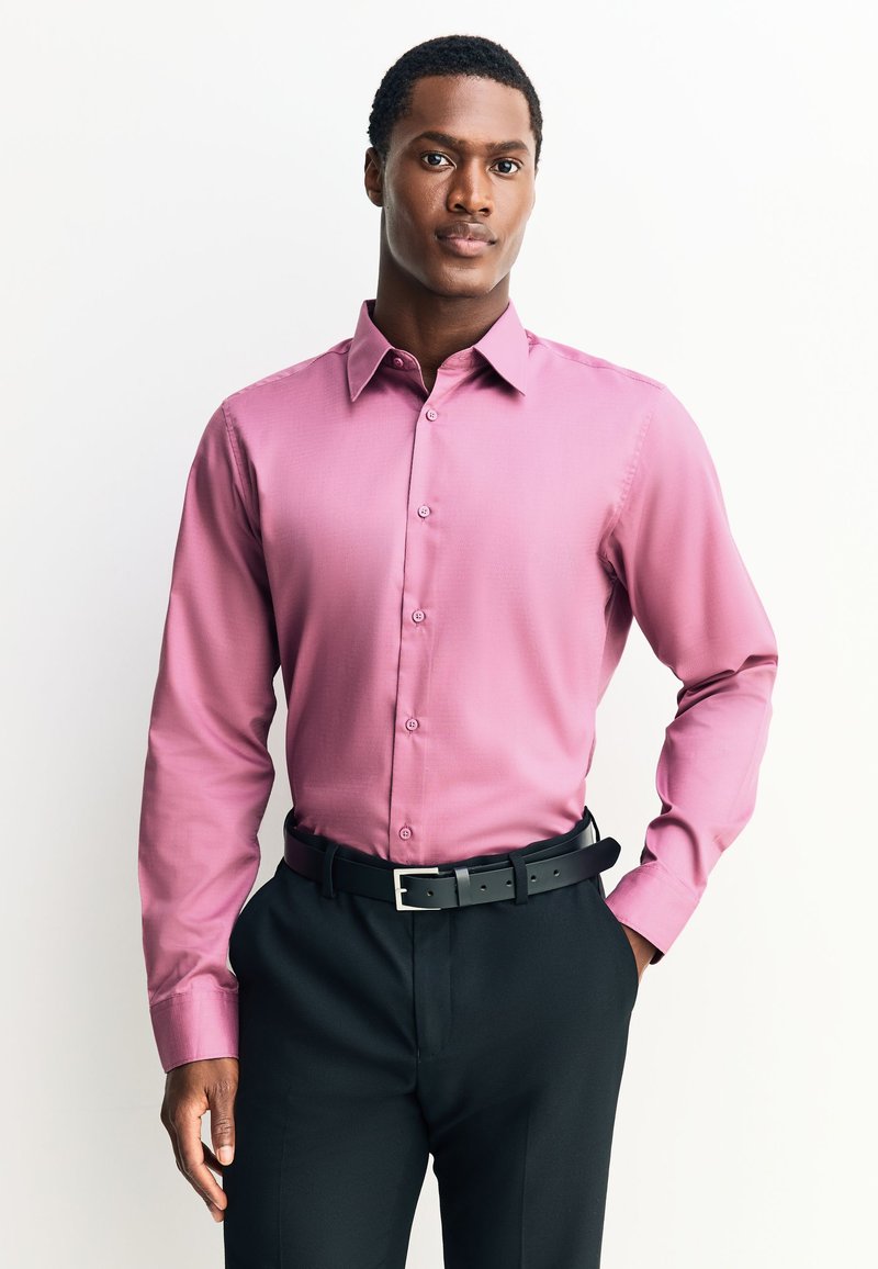 Różowa koszula wizytowa z długim rękawem, z ostrym kołnierzykiem, zapinana na guziki z przodu, wykonana z teksturowanej tkaniny, zestawiona z czarnymi spodniami i paskiem.