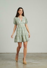 Vestido floral verde claro con mangas abullonadas y escote en V; dobladillo con volantes, patrón similar al encaje. Llevado con tacones de plataforma color beige. Textura suave.
