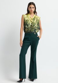 Mouwloos topje in geel met groene bloemenprint; hoogsluitende groene broek. Opvallende gouden hardware-accent op de tailleband.
