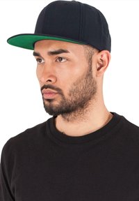 Fekete baseball sapka zöld karimával, strukturált dizájn, és lapos csúccsal. Rövid hajú és szakállas férfi viseli, alatta fekete pólóval.