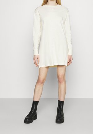 Femme portant une robe Nike à manches longues de couleur ivoire associée à des bottes de combat noires à lacets, debout devant un fond uni.