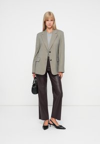 Szürke kockás zakó két gombbal, barna műbőr nadrággal és fekete hegyes orrú magassarkú cipővel párosítva, egy fekete kézitáskát tartva.