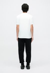 Λευκό κοντομάνικο μπλουζάκι, μείγμα βαμβακιού, άνετη εφαρμογή, σε συνδυασμό με μαύρα παντελόνια jogging και μαύρα παπούτσια, από πίσω σε απλό φόντο.