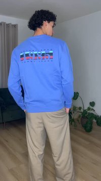 Mees lokkis juustega, kes kannab sinist pusa, mille seljal on kiri "1985 Tommy Jeans", beežide pükstega, seisab siseruumis taimedest ja kardinast lähedal.