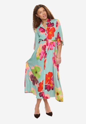 Kvinde iført en turkis knapbesat kjole med store farverige blomsterprint, der holder nederdelen og står i sorte spidse hæle.