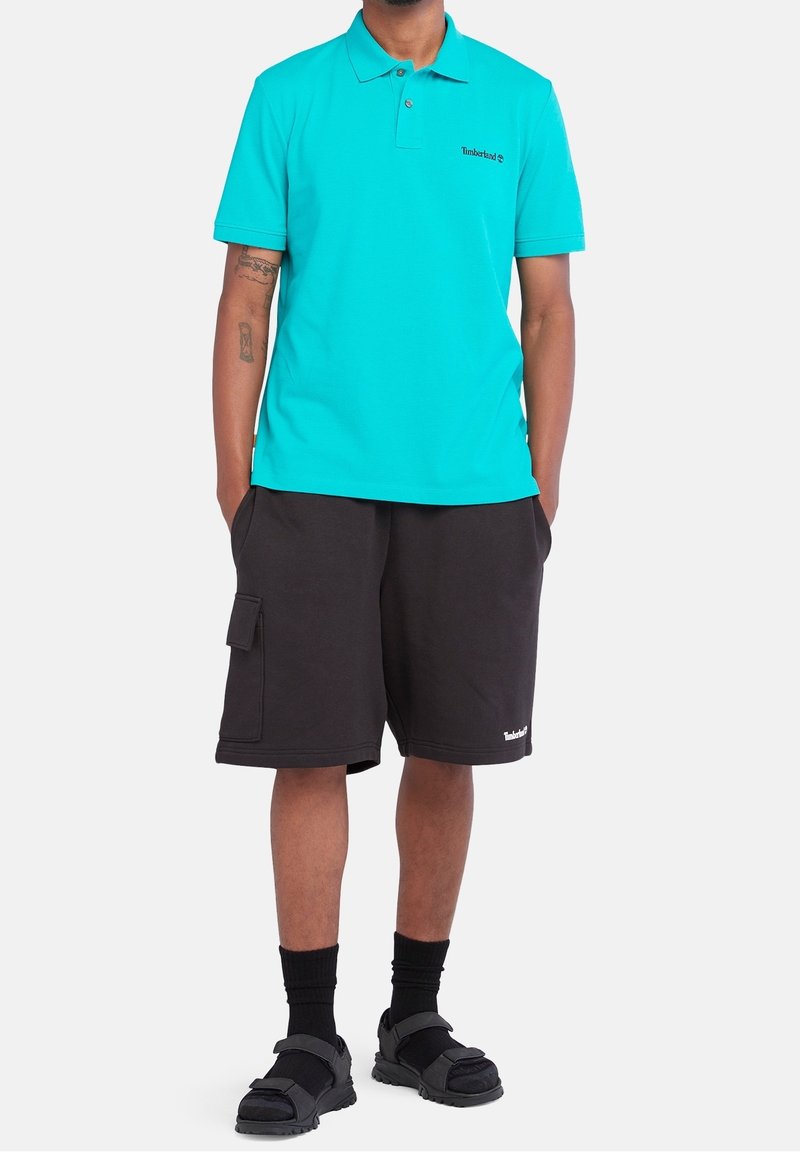 Homme portant un polo turquoise, un short cargo noir, des chaussettes noires et des sandales noires, debout les mains dans les poches.