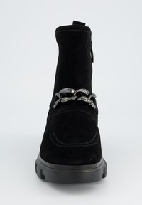 Bottine en daim noir avec fermeture éclair sur le côté, caractérisée par une semelle plate-forme noire épaisse et une chaîne métallique décorative sur l'avant.