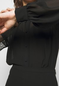 Schwarze durchsichtige Bluse mit voluminösen Ärmeln, ausgestattet mit Knopfverschluss und einer glatten Textur. Das Design der Bluse umfasst eine lockere Passform.