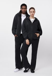 Δύο άτομα φοράνε ταιριαστά μαύρα βαμβακερά φούτερ με κουκούλα, εμπρόσθια φερμουάρ και τσέπες, συνδυασμένα με μαύρο φόρμα παντελόνι και διάφορα αθλητικά παπούτσια.