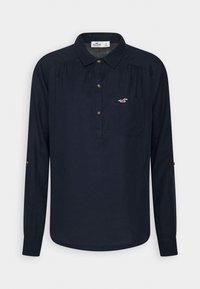 Chemise bleu marine à manches longues avec col, patte à trois boutons, poche poitrine et petit logo oiseau blanc et rouge sur la poche.
