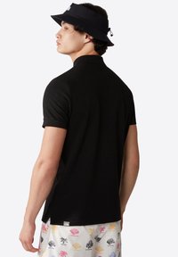 Czarny T-shirt polo z krótkimi rękawami, teksturowana tkanina i subtelna metka z logo na dole. Noszony z szortami w wzory z kolorowymi motywami.