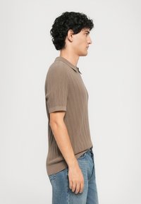 Ung mann som står sidelengs iført en brun kortermet strikket piké skjorte og blå jeans mot en ensfarget bakgrunn.