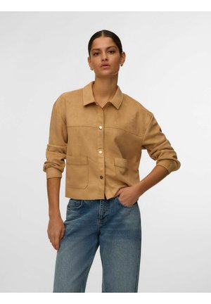 Žena u smeđoj jakni na kopčanje s dva prednja džepa i plavim trapericama, stoji s jednom rukom u džepu ispred jednobojne pozadine.