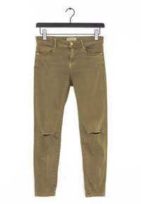 Jeans ajustados de color verde oliva con desgarros desgastados en las rodillas, diseño de cinco bolsillos y cremallera, colgando de una percha negra.