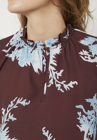 Mørkerød bluse med lys blå blomsterprint, med en plisseret halsudskæring og en knaplukning foran. Glat stofstruktur.