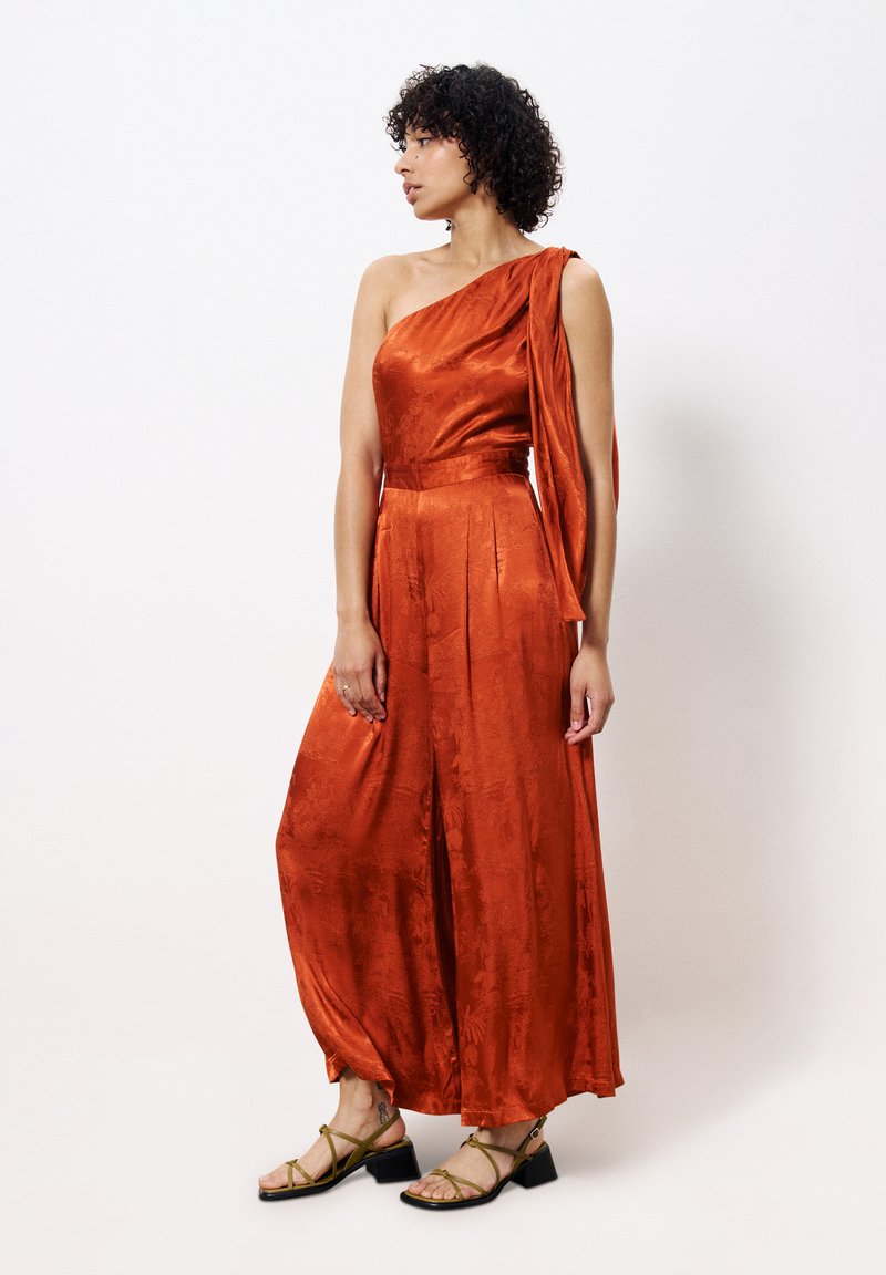 Een off-shoulder jumpsuit in glanzend oranje stof met een gedrapeerd detail en een wijde pijpen silhouette. Gepaard met groene sandalen met bandjes.