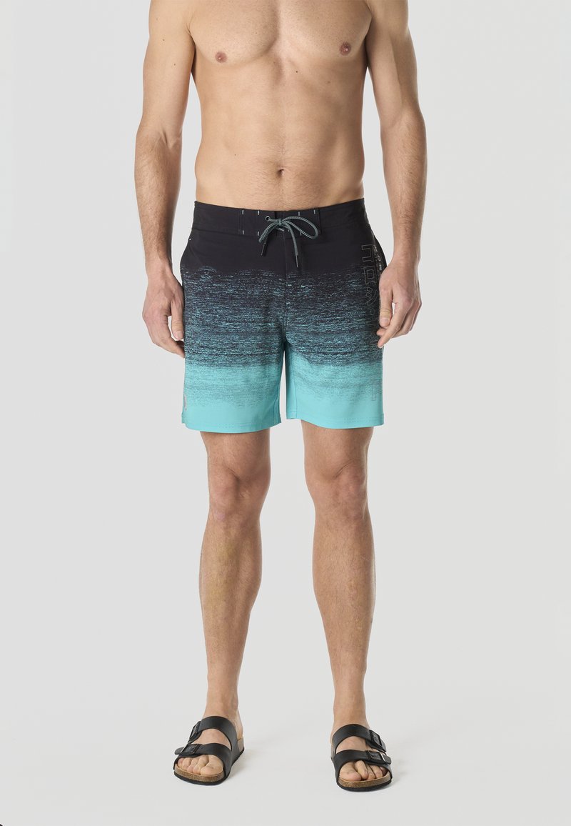 Torso e gambe maschili che indossano pantaloncini da bagno sfumati neri e turchesi e sandali neri, in piedi contro uno sfondo chiaro e uniforme.