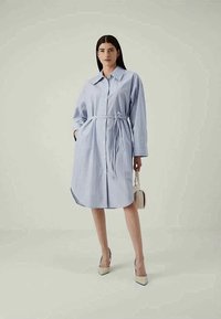 Γυναικα στέκεται φορώντας ένα ανοιχτό μπλε ριγέ φόρεμα με ζώνη στη μέση, κρατώντας μια μικρή μπεζ τσάντα και φορώντας μπεζ μυτερές γόβες.