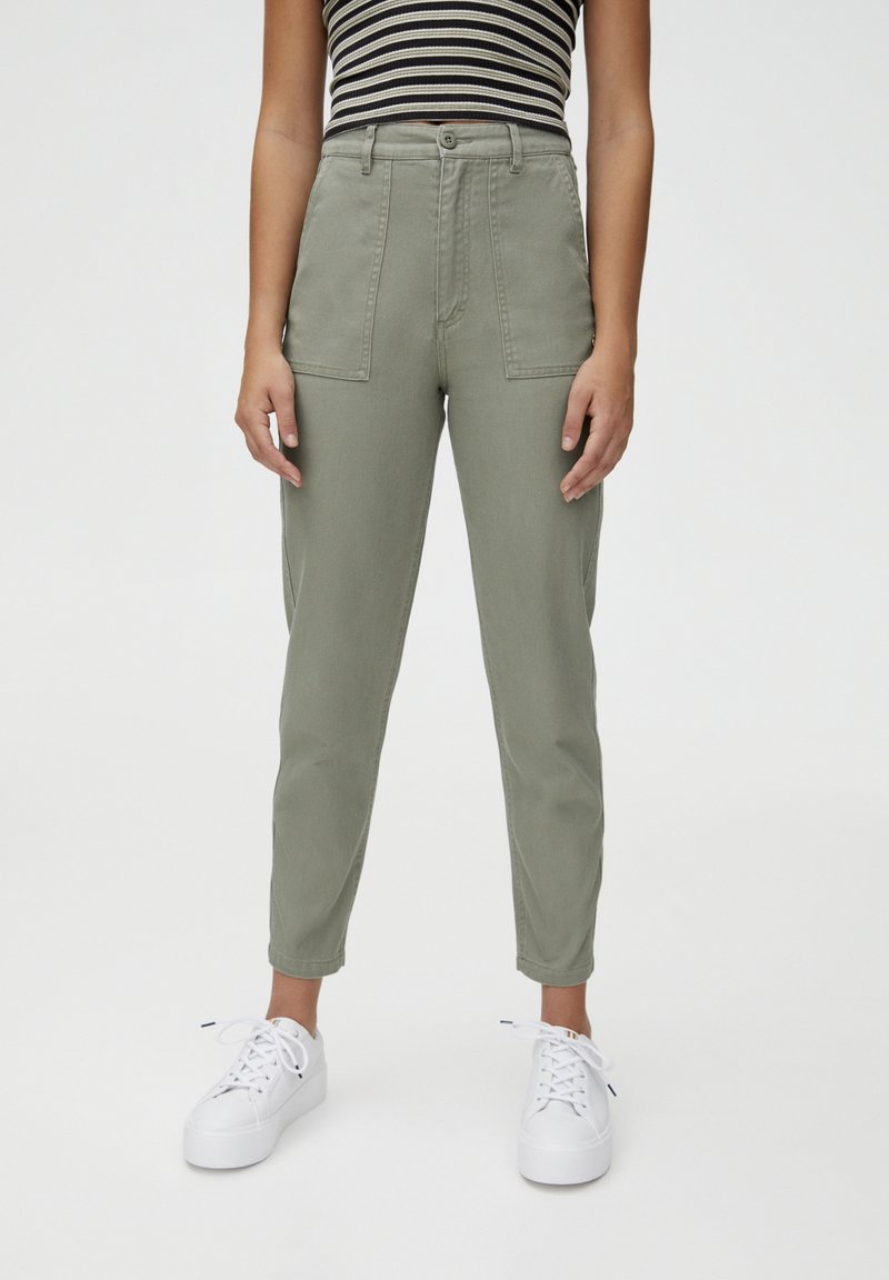 Femme portant un pantalon taille haute vert olive avec des poches plaquées à l'avant, un top rayé noir et blanc sans manches, et des baskets blanches.