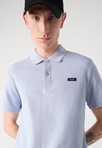 Polo bleu clair en tissu texturé, à col classique, avec deux boutons et une étiquette de marque noire sur la poitrine. Logo discret sur le col.