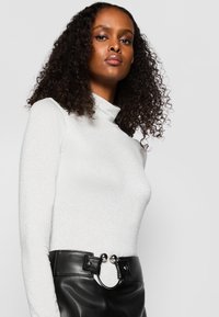 Weißes Oberteil mit langen Ärmeln und hohem Kragen aus schimmerndem Stoff; kombiniert mit einem schwarzen Lederrock, der eine einzigartige kreisförmige Gürtelschnalle hat.