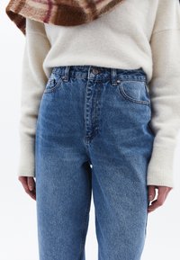 Höga blå jeans med en blekt finish, femficksdesign och kopparfärgad knapp; bärs med en krämfärgad tröja.