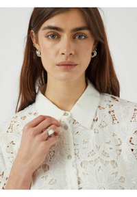 Vit spetsblus med krage och knäppning, med intrikata blommönster och texturerade detaljer. Modell som bär en silvring.
