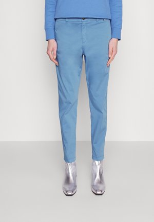 Persona vistiendo pantalones ajustados azul claro, camiseta de manga larga azul y botines plateados brillantes, de pie contra un fondo liso.