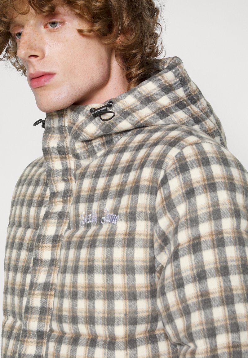 Vadderad jacka i ett rutmönster av beige, grå och brun. Har en hög krage, dragkedjestängning och en broderad logotyp på bröstet.