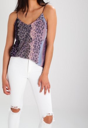 Woman wearing a sleeveless patterned top with thin straps and white ripped skinny jeans, standing against a plain light background.