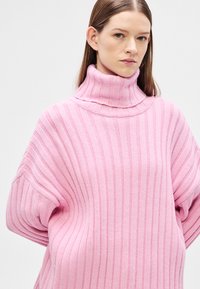 Růžový pletený rolák s vertikálním žebrováním, volným střihem a širokými rukávy. Šev je zdůrazněn u výstřihu.