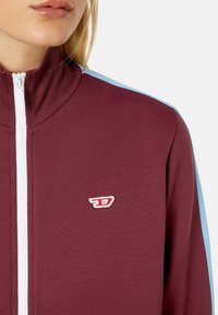 Burgundská zipová bunda s vysokým límcem, s světle modrými pruhy na ramenech a malým červeným logem na hrudi. Hladká struktura tkaniny.