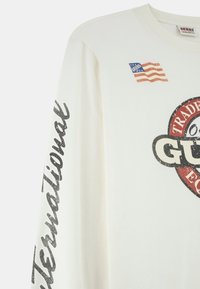 Witte langwerpige t-shirt met lange mouwen, een ronde grafische logo, glanzende zwarte tekst op de mouw en een kleine Amerikaanse vlag op de voorkant.