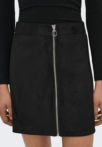 Svart semsket miniskjørt med en frontmetallglidelås, med en glatt tekstur og minimalistisk design. Uthevet med en sølvglidelåstrekk.