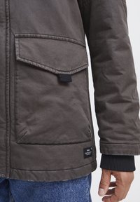 Chaqueta acolchada gris con un gran bolsillo frontal, puños acanalados y una pequeña etiqueta de marca cerca del dobladillo. La textura de la tela es duradera y suave.