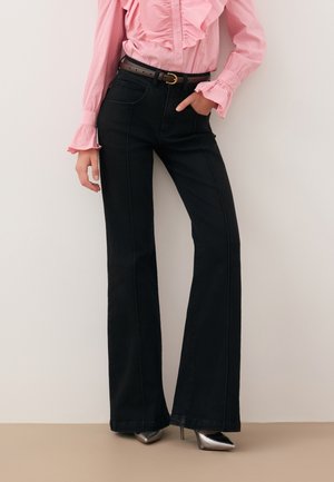 Vrouw draagt zwarte flare broek met hoge taille, bruine riem, roze blouse met ruches en zilveren puntige hoge hakken, staand tegen een beige muur.