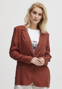Prevešena jakna iz rdeče barve rjave boje z izrezom, dvema sprednjima žeparjema in ohlapnim krojem, preplastena preko bele majice s potiskom.