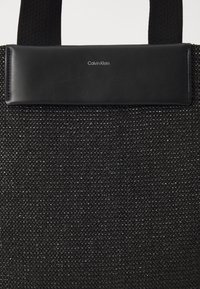 Czarna torba tote z teksturowanej tkaniny z wyściełanym skórzanym uchwytem, na którym znajduje się biały logo Calvin Klein umieszczone centralnie.