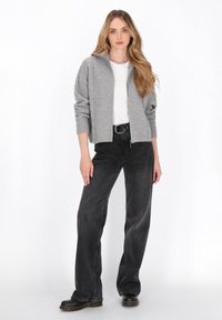 Cardigan gri cu fermoar, cu accente din tricot cu cablu, purtat peste un tricou alb. Jeanși negri cu picioare largi și saboți negri completează ținuta.