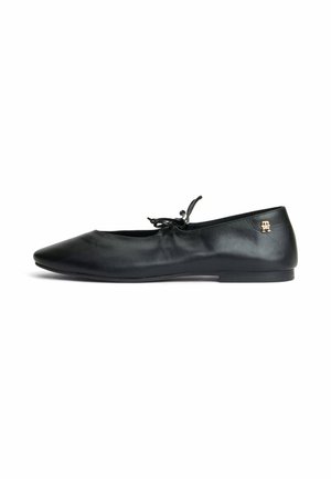 Schwarzer Leder-Ballettschuh mit einer kleinen Schleife oben und einem goldenen Logo-Emblem nahe der Ferse auf weißem Hintergrund.