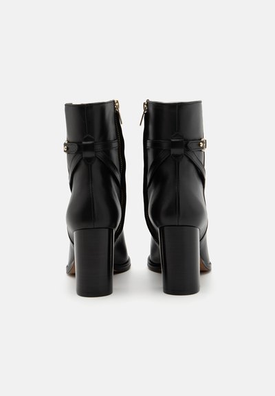 Bottines en cuir noir avec fermeture éclair, présentant un design croisé, une sangle décorative et des talons bloc robustes. Texture lisse tout au long.