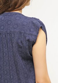 Persona vistiendo una blusa sin mangas de color azul marino con un sutil patrón floral circular, vista desde la parte de atrás y el área del hombro.