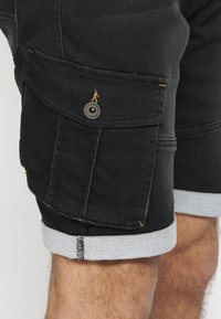 Shorts de mezclilla negra con un dobladillo en gris claro, bolsillos laterales y detalles de costura amarilla. Se ve un acento de botón metálico circular.