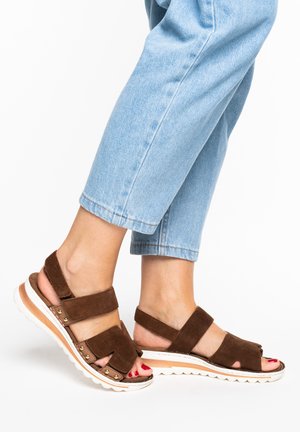 Piedi che indossano sandali in camoscio marrone con suole bianche e borchie dorate, abbinati a jeans azzurri corti su sfondo bianco.